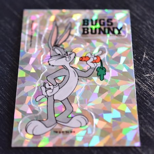 May include: A holographic sticker featuring Bugs Bunny holding a carrot. The sticker has a white background with a holographic pattern and the text "Bugs Bunny" in black.