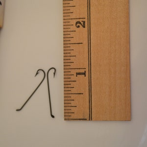 May include: Two black metal hooks, each about 1 inch long, are placed on a white surface next to a wooden ruler marked with inches. The ruler is marked with numbers 1, 2, and 3.