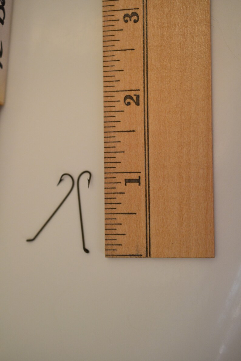 May include: Two black metal hooks, each about 1 inch long, are placed on a white surface next to a wooden ruler marked with inches. The ruler is marked with numbers 1, 2, and 3.