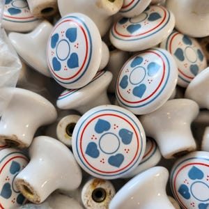 May include: A selection of white ceramic cabinet knobs. Each knob has a circular design with blue hearts, a red and blue border, and small red dots. The knobs are rounded and ready for installation.