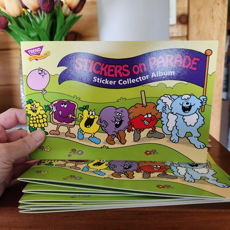 May include: A stack of "Stickers on Parade" sticker collector albums. The cover features a green background with cartoon characters marching across a path. The album's title is in purple and white text. A hand is holding the top album.