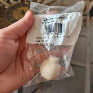 May include: A close-up of a white plastic cabinet knob with a screw. The knob is packaged in a clear plastic bag with a label that reads "Amerock The Newton Group BP727A-AM 0 26634 10140 1 Rockford, IL. U.S.A. 61125 798 MADE IN USA FABRIQUE AU USA".