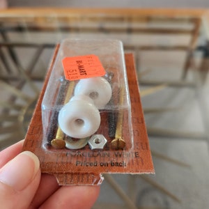 Peut inclure: Deux boutons de cabinet en porcelaine blanche avec des vis dorées et un écrou argenté dans un emballage en plastique transparent. L'emballage porte une étiquette de prix rouge et blanche indiquant "BI-MART" et "1.57 EACH".