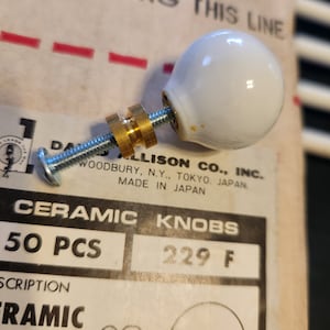 May include: A package of 50 white ceramic knobs with a gold screw and a silver base. The package says "Ceramic Knobs 50 PCS 229 F" and "Allison Co., Inc. Woodbury, N.Y., Tokyo, Japan. Made in Japan."