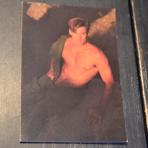May include: A shirtless man with short brown hair sits on a rock with a green shirt draped over his shoulders. The background is a dark, blurry forest.