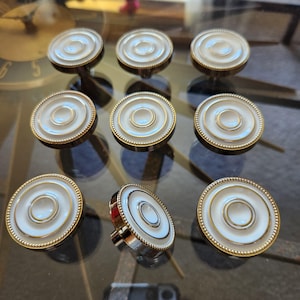 May include: Nine decorative cabinet knobs with a white and gold design. Each knob features a circular face with concentric rings of white and gold, and a gold-coloured base. The knobs are arranged on a reflective surface.