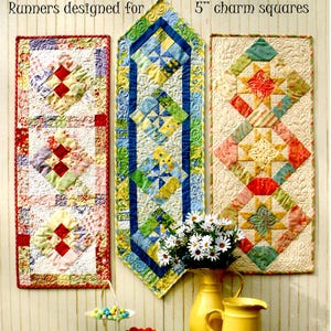 May include: Three colorful table runners made from 5 inch charm squares. The runners are displayed on a white wall with a vase of white flowers in front of them. The runners are made in a variety of patterns and colors, including red, blue, yellow, green, and pink.