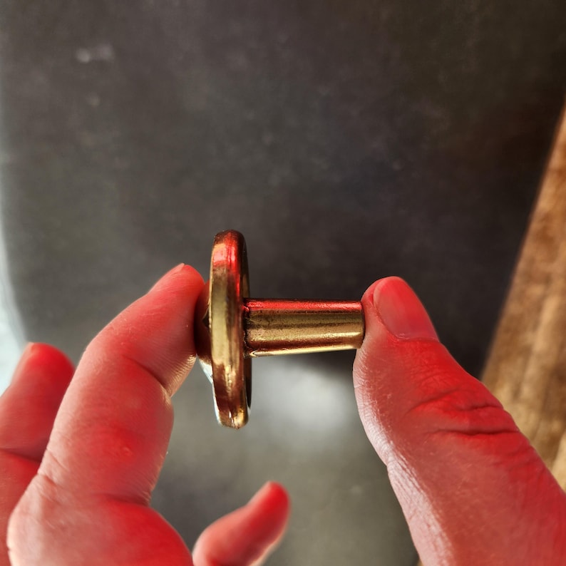 May include: A gold-colored metal knob with a round base and a cylindrical stem. The knob is held in a hand.