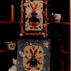 May include: Two framed quilted wall hangings featuring a bunny design with buttons and bows. The top hanging has a pink and brown bunny with a blue dress and the bottom hanging has a blue and brown bunny with a blue dress. The hangings are on a shelf with other decorative items.