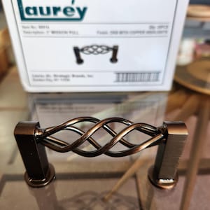 May include: A bronze-colored cabinet pull with a decorative woven design. The pull has a rectangular base and a curved, intertwined handle. The packaging label reads "1" MISSION PULL".
