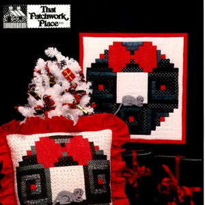 Puede incluir: Una almohada de patchwork roja y negra con fondo blanco y un pequeño ratón gris en el centro. La almohada tiene un volante rojo alrededor del borde. La almohada está sentada en una mesa con un árbol de Navidad blanco y una corona de patchwork roja y negra en el fondo. El texto "That Patchwork Place" es visible en la esquina superior izquierda de la imagen.