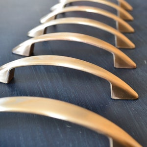 May include: A row of eight brushed nickel cabinet pulls, arranged in a staggered pattern on a dark blue surface. The pulls are all the same size and shape, with a slightly curved design.