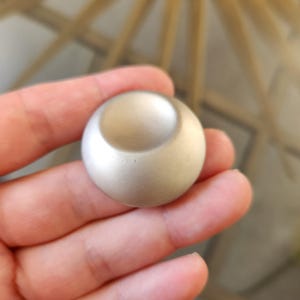 May include: A small, silver-colored, spherical object with a curved indentation on top. The object is smooth and appears to be made of metal. The design is minimalist and modern. The object is held in a hand.