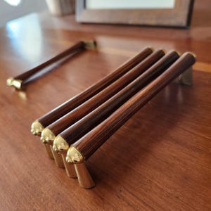 May include: A set of four drawer pulls featuring dark brown wood grain cylindrical bars with polished gold-tone metal end caps and mounting brackets. Each pull is arranged in a parallel row, with one additional matching pull placed separately.