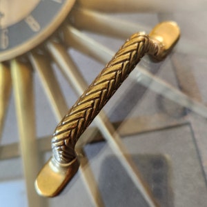May include: A gold-toned, decorative cabinet pull with a woven design. The pull is shaped like a long, thin handle with a rounded end.
