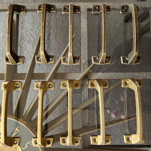 May include: Eight gold-toned metal cabinet pulls with a rectangular shape and a slightly curved handle. The pulls have a brushed finish and are arranged in two rows of four.