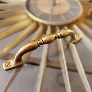 May include: Close-up of a gold-toned clock with a sunburst design. The clock face is dark with Roman numerals. A curved, brass handle is in the foreground. The clock's design is mid-century modern.