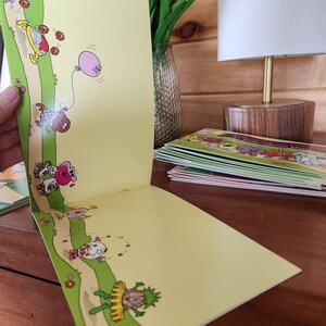 May include: Open book with colorful illustrations of whimsical characters on a yellow background. The illustrations feature a path with characters, a pink balloon, and various objects. A stack of similar books is visible in the background.