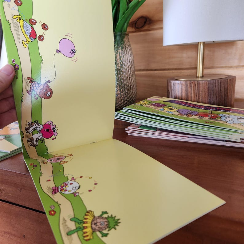 May include: Open book with colorful illustrations of whimsical characters on a yellow background. The illustrations feature a path with characters, a pink balloon, and various objects. A stack of similar books is visible in the background.