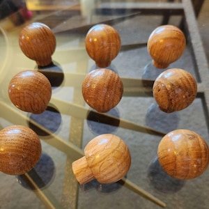 May include: Nine wooden cabinet knobs with a natural wood grain finish. The knobs are spherical with a short, cylindrical base. The wood has a warm, light brown color, and the grain is visible. The knobs are arranged on a reflective surface.