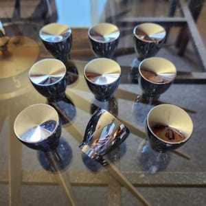 May include: A collection of nine shiny, chrome-finished knobs with a rounded, reflective top and a dark, cylindrical base. The knobs are arranged on a glass surface, reflecting the surrounding environment.