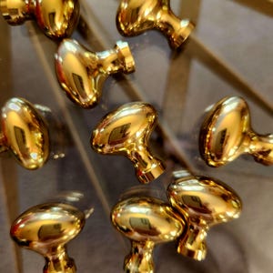 May include: A collection of polished gold-tone metal cabinet knobs, each featuring a smooth, rounded mushroom-shaped head and a tapered cylindrical base. The metallic surface is highly reflective, showing clear highlights and surrounding light.