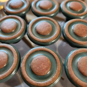 May include: A collection of nine circular cabinet knobs arranged in a grid. Each knob features a central raised brown disc surrounded by a smooth, dark teal ring. The surface of the brown sections shows a subtle, cracked, and textured finish.