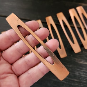 May include: Copper-colored hair clips with an oval cutout in the center. The clips have a rectangular shape with rounded edges. The metal has a slightly aged appearance. Several clips are visible, some in focus, others blurred.