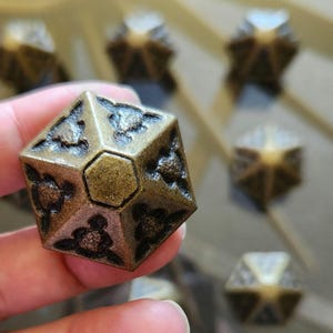 May include: A bronze-colored, multi-sided die with a geometric design. The die has a series of turtle-like shapes on each side. Other dice are visible in the background, slightly out of focus.