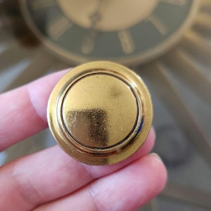 May include: A gold-coloured, circular button with a raised outer rim and a flat, central disc. The button has a shiny, metallic finish. The background shows a portion of a clock.