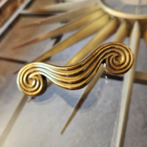 May include: A gold-toned metal cabinet handle featuring a wave-like design with prominent spiral curls at both ends. The surface has a polished finish with subtle dark highlights in the recessed grooves, creating a textured appearance.