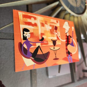 May include: A framed art print with a stylized illustration of two figures in a room. The woman, in purple, sits in a chair with a black bird. The man holds a book. The background is orange with abstract shapes.