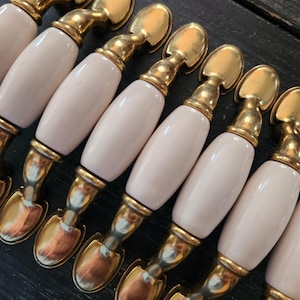 May include: A collection of cabinet pulls with a light pink, oval-shaped ceramic handle and gold-toned metal hardware. The pulls are arranged in a row, showcasing their decorative design and potential for furniture upgrades.