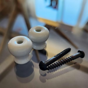 May include: Two white ceramic cabinet knobs with a smooth, rounded shape and a central hole, accompanied by two long, dark metal screws with threaded shafts and rounded heads, designed for installation on furniture or cabinetry hardware.