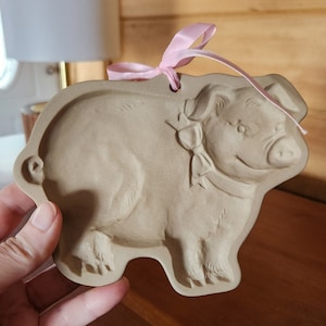 May include: A tan-colored pig-shaped ornament with a pink ribbon for hanging. The pig has a smiling face and a bow. The ornament is made of a ceramic material. The background is a blurred interior setting.