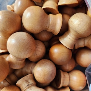 May include: A collection of unfinished wooden knobs with a rounded top and a flared base. The knobs are a light brown color and appear to be made of natural wood. The image shows a close-up view of the knobs, with many of them overlapping.