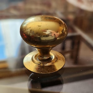May include: A vintage brass doorknob with a round, ball-shaped handle and a circular base. The brass has a warm, golden hue with signs of wear, suggesting age. The doorknob is set against a reflective surface.