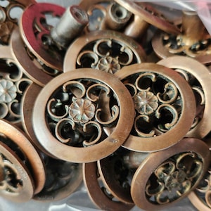May include: A collection of antique bronze-colored drawer pulls. Each pull features a circular design with an ornate floral center and swirling patterns. The hardware is likely for furniture restoration or vintage decor.