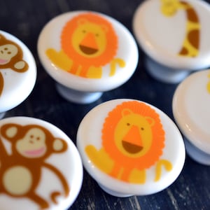 May include: Six white ceramic knobs with different animal designs. The knobs feature a monkey, a lion, and a giraffe. The animals are depicted in a cartoon style with bright colours.