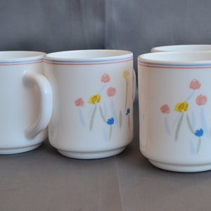 May include: Set of four white ceramic mugs with a floral design in shades of blue, pink, yellow, and green. The mugs have a simple, rounded shape and a comfortable handle.