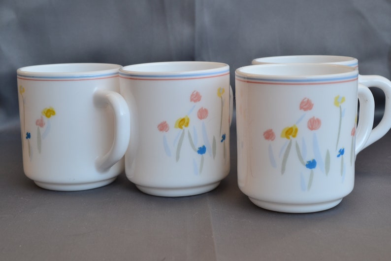 May include: Set of four white ceramic mugs with a floral design in shades of blue, pink, yellow, and green. The mugs have a simple, rounded shape and a comfortable handle.