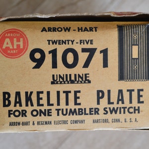 May include: A brown cardboard box with the text "ARROW-HART TWENTY-FIVE 91071 UNILINE TRADE MARK BAKELITE PLATE FOR ONE TUMBLER SWITCH ARROW-HART & HEGEMAN ELECTRIC COMPANY HARTFORD, CONN., U. S. A." printed on it. The box has a red circle with the letters "AH" in white inside the circle and the word "HART" below the letters. The box also has a black and white illustration of a light switch plate.