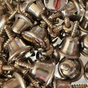 May include: Close-up of a pile of small, shiny silver bells with screw-like stems. The bells have a classic bell shape with a smooth, reflective surface. The image is a close-up, showing the details of the metal and the screw threads.