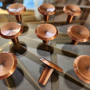 Peut inclure: Ensemble de boutons de meubles de couleur cuivre avec une finition brossée. Les boutons ont un dessus plat et arrondi et une tige cylindrique. La quincaillerie est présentée sur une surface réfléchissante, mettant en valeur les tons chauds et la texture métallique.