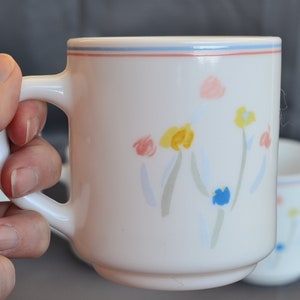May include: White ceramic mug with a blue, pink, and yellow floral design. The mug has a handle and a blue and red rim.