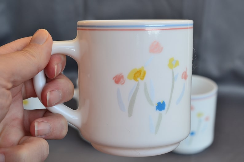 May include: White ceramic mug with a blue, pink, and yellow floral design. The mug has a handle and a blue and red rim.
