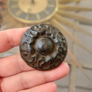May include: A dark brown metal knob with a raised, circular design. The knob has a textured surface and a slightly raised center.