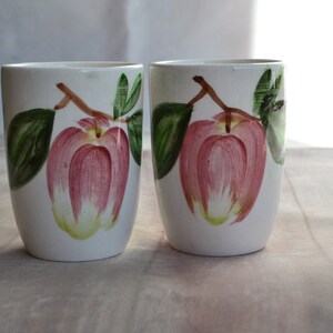May include: Two white ceramic mugs with a hand-painted design of pink apples and green leaves.