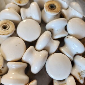 May include: A collection of white ceramic cabinet knobs with a smooth, rounded top and a flared base. Each knob features a central metal threaded insert on the underside, showing signs of age and wear with some brown discoloration on the base.
