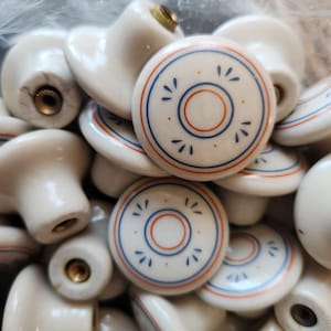 May include: A collection of round white ceramic cabinet knobs featuring a decorative pattern of concentric blue and orange rings with small blue petal-like accents. Each knob has a central brass-coloured metal threaded insert on the back.